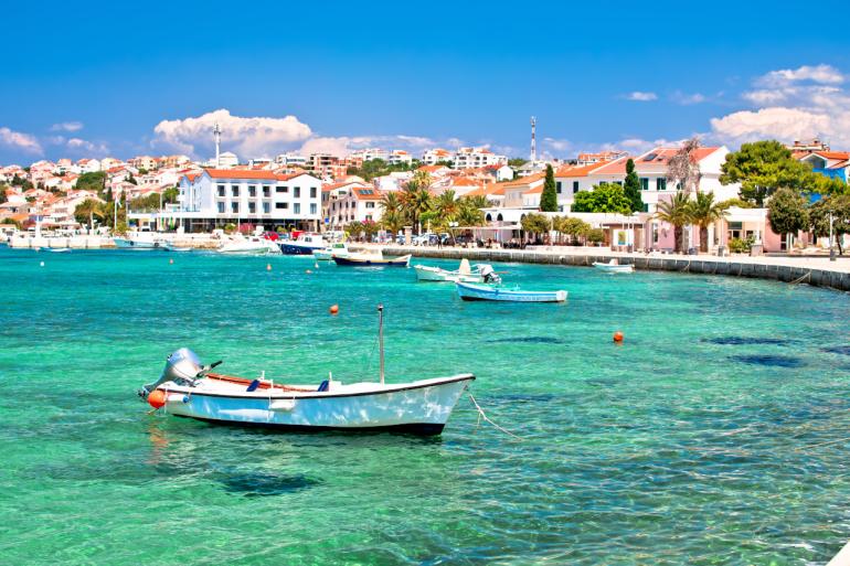 Widok na nadmorskie miasteczko Pag w Chorwacji. W krystalicznie czystej, turkusowej wodzie Adriatyku unoszą się białe łodzie i jachty. W tle widać promenadę z palmami, pastelowe budynki z czerwonymi dachami oraz niewielkie hotele i restauracje położone tuż przy brzegu.