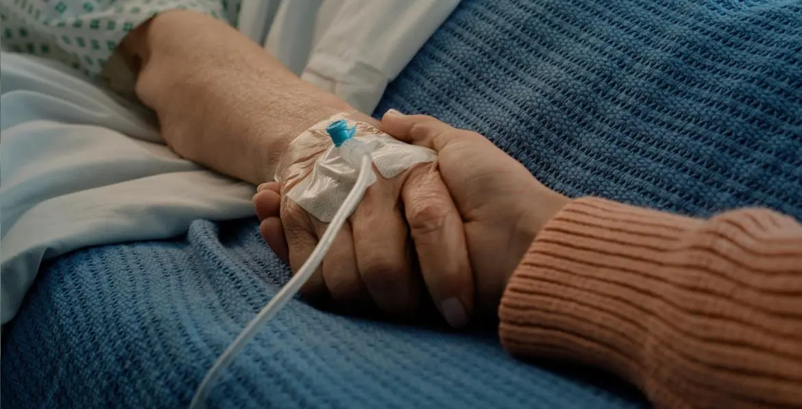 dłoń trzymająca dłoń chorej osoby z wenflonem i kroplówką
