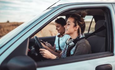 Dwie młode dziewczyny w białym samochodzie. Jedna z nich uśmiecha się i siedzi za kierownicą. Druga patrzy w telefon