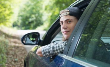 Młody chłopak za kierownicą samochodu. Ma czapkę z daszkiem, patrzy w obiektyw i ma wystawiony łokieć za szybę