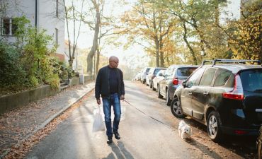 Mężczyzna spaceruje z psem. W ręce niesie siatkę z zakupami. Obok stoją zaparkowane na chodniku samochody.