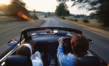 Mężczyzna prowadzi samochód. Obok niego siedzi kobieta. Jadą szybko lewym pasem. Samochód ma odsłonięty dach.