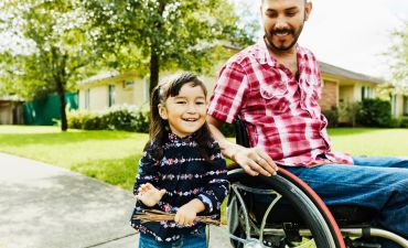 Mężczyzna siedzi na wózku inwalidzkim. Obok niego stoi dziewczynka i się uśmiecha